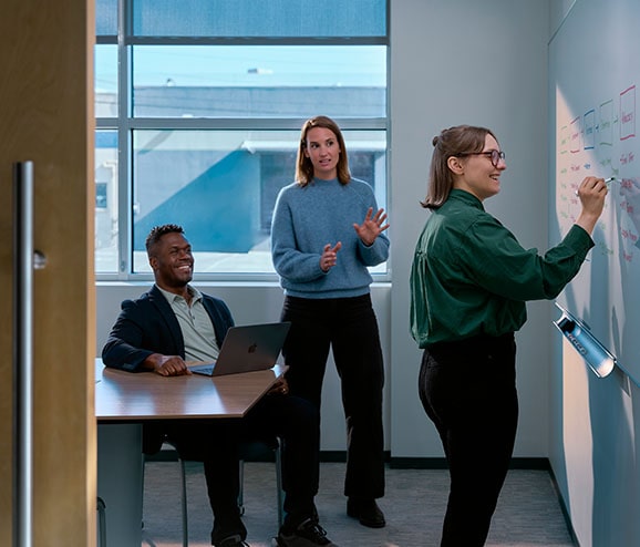 A white woman charts a customer journey on a whiteboard while a white woman in a blue sweater and a Black man in a suit confer at a table.