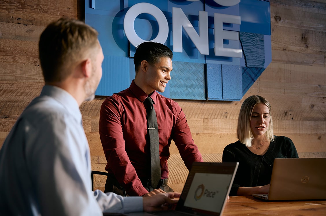 An Asian man, white woman, and white man compare work over their laptops. A blue DCG ONE screen hangs on a wood-paneled wall behind them.
