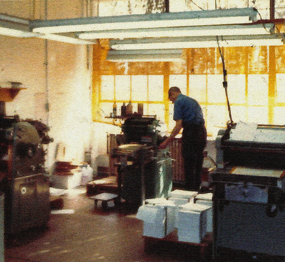 Blurry photo of a man in an industrial room working one of several printers, reams of paper stacked around him.